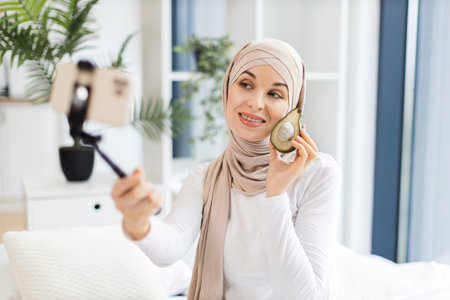 Young Muslim woman in beige hijab holding smartphone on tripod sharing skincare tips using avocado. She discusses natural beauty methods while creating engaging and insightful content in a cozy homeの写真素材