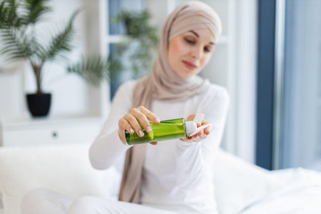 Young Muslim woman wearing hijab applying lotion for skincare in bright modern room. Focusing on...