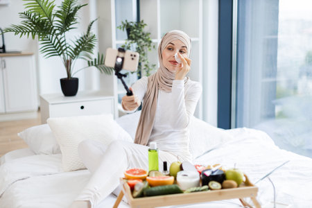 Muslim woman in loungewear using cotton pads and skincare products while recording self-care blog in her modern, bright home interior. Captures contemporary beauty routine and home lifestyle.の写真素材