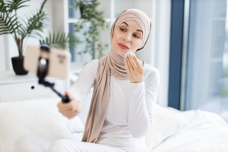 Young Muslim woman in light-colored hijab using cotton pads, blogging about skincare routine and personal health, sitting in an elegant and sunlit room while capturing content on a mobile device.の写真素材