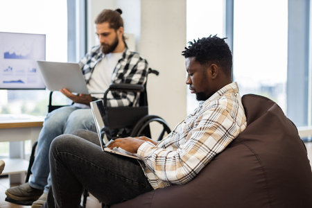Coworkers, one seated in wheelchair and other on beanbag, working with laptops, showcasing...