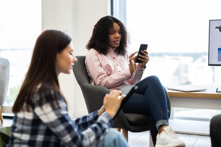 Two young adult women in casual clothing using digital devices in a bright office. One woman using...