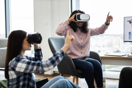 Women interacting with virtual reality technology wearing VR glasses in office setting. Exploring...