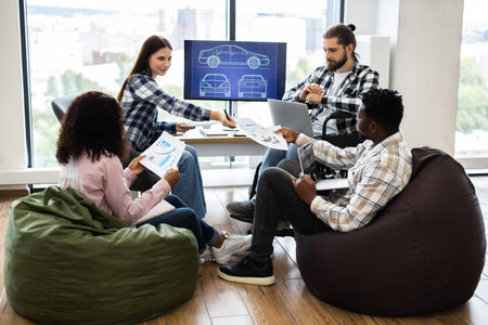 Young diverse adults discussing car design concepts in office using documents and screens. Collaborative effort with focus on teamwork and innovative solutions for engineering and construction tasksの写真素材