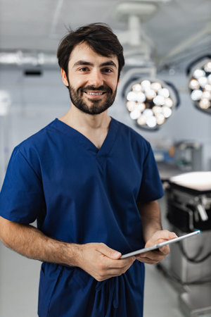 A smiling surgeon in scrubs holds a tablet in an operating room, illuminated by surgical lights.の写真素材