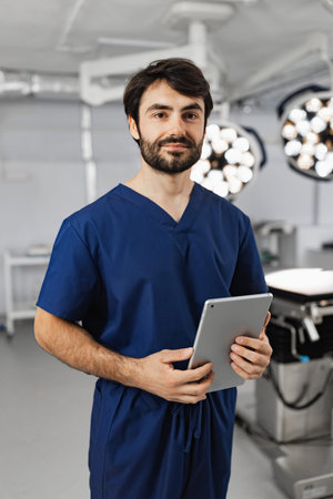 A surgeon in scrubs stands in an operating room, holding a tablet and looking at the camera.