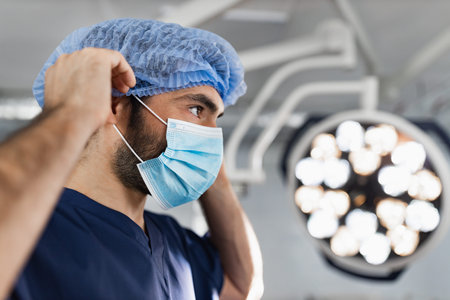 A surgeon adjusts his surgical cap and mask in a modern operating room, preparing for a procedure.