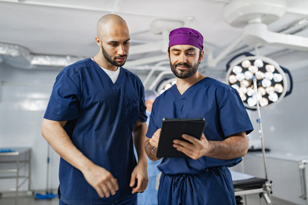 Two surgeons in scrubs review a tablet in an operating room, discussing patient information before a...