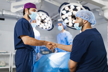 Two surgeons shake hands in an operating room, with other medical staff and surgical equipment in...
