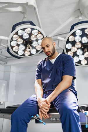 A surgeon takes a moment to rest in an operating room, illuminated by surgical lights.