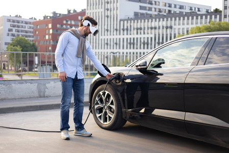 A man wearing VR glasses plugs an electric vehicle into a charging station in an urban setting.の写真素材