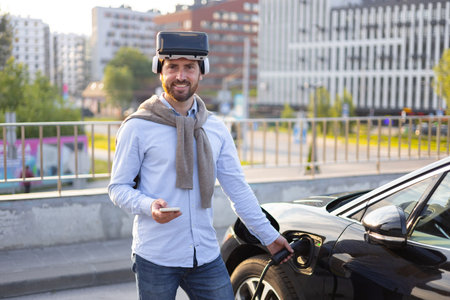 A man wearing a VR headset smiles while charging his electric car at a charging station in an urban...