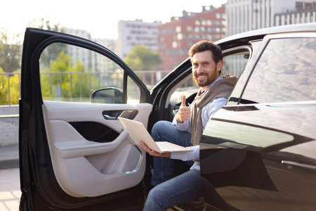 A smiling man gives a thumbs up while sitting in a car with a laptop, suggesting success or...