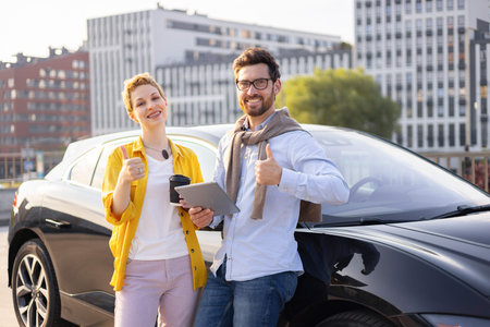 A smiling couple stands next to a black electric car, giving thumbs up, with a tablet and coffee...