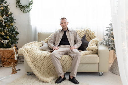 A man sits on a comfortable couch in a bright, cozy living room, surrounded by Christmas decorations.の写真素材