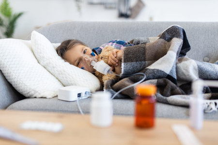 A young child lies on a couch, using a nebulizer to help with breathing while holding a teddy bear,...