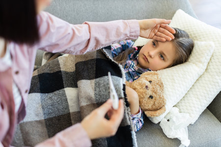 A concerned parent checks the temperature of a sick child with a thermometer while the child rests on a couch with a teddy bear.の写真素材