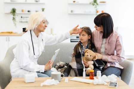 A doctor checks a young girls temperature while the mother comforts her, suggesting a possible illness or fever. The scene takes place in a home setting.の写真素材