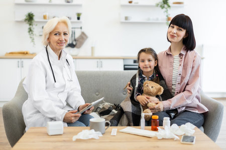 A doctor is consulting with a mother and her sick child, who is wrapped in a blanket and holding a teddy bear, at home.の写真素材