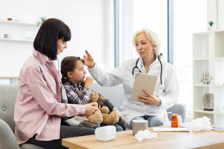 A doctor checks a young girls temperature while her mother comforts her in a doctors office, with a teddy bear.の写真素材
