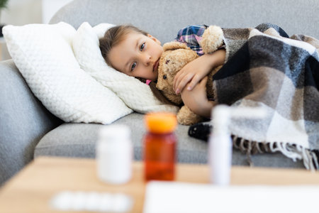 A young girl, wrapped in a blanket, lies on a sofa with a teddy bear, appearing unwell. Medication bottles sit on a table in the foreground.の写真素材