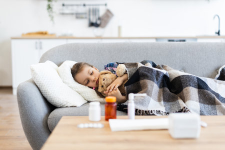 A young girl sleeps soundly under a blanket, clutching a teddy bear, with medicine on the table nearby, suggesting illness.の写真素材