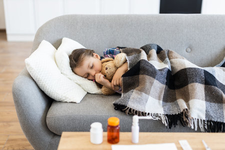 A young girl rests on a sofa, cuddled under a blanket with a teddy bear, while medicine sits nearby, suggesting illness.の写真素材