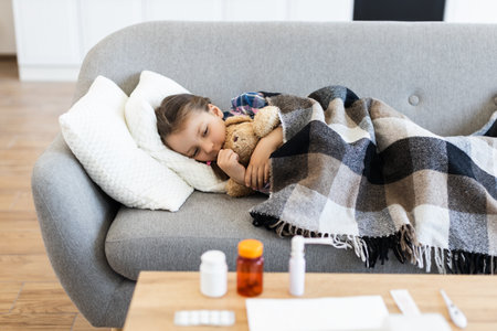 A young girl, wrapped in a blanket, is sleeping on a sofa. Various medicines are placed on a table in front of her, suggesting illness.の写真素材