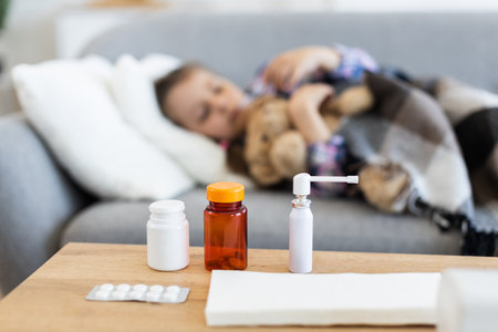 A young child rests on a sofa, appearing unwell, with various medications placed on a table in the foreground, suggesting illness.の写真素材