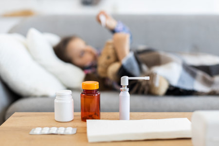 A young girl rests on a couch, appearing ill, with medicine and tissues on a nearby table, suggesting illness.の写真素材