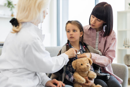 A doctor uses a stethoscope to examine a young girl while her mother comforts her, teddy bear in hand, during a checkup.の写真素材