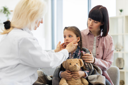 A doctor checks a young girls throat while her mother comforts her. The girl is wrapped in a blanket, holding a teddy bear.の写真素材