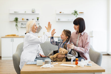 A doctor is giving a high-five to a young girl, who is sitting with her mother on a couch. The girl is holding a teddy bear, and there are medicine bottles on the table.の写真素材