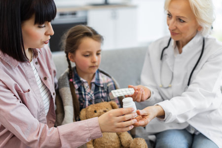 A doctor is giving medicine to a sick child, with the mother present, likely at a home visit. The child is wrapped in a blanket and has a teddy bear.の写真素材