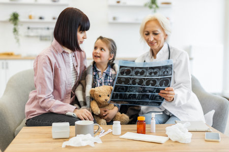 A doctor explains an X-ray to a mother and her young daughter, who is holding a teddy bear, in a cozy setting.の写真素材