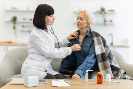 A female doctor is using a stethoscope to listen to an elderly womans heart and lungs during a home visit. Various medicines are on the table.の写真素材