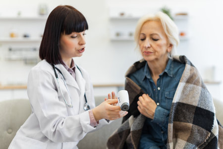 A female doctor in a white coat checks an elderly woman's temperature with a digital thermometer. The patient is wrapped in a blanket.の写真素材