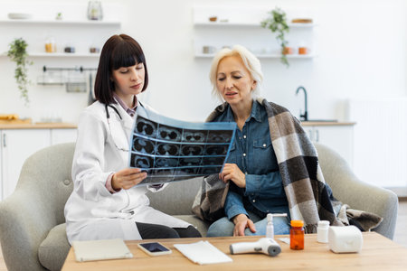 A female doctor reviews an X-ray with an elderly woman wrapped in a blanket on a sofa, discussing the results in a home setting.の写真素材