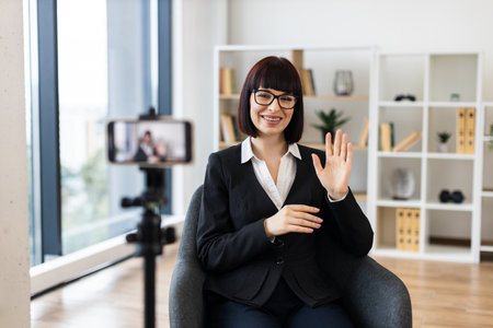 Caucasian adult woman in formal attire recording business blog using smartphone on tripod indoors.の写真素材