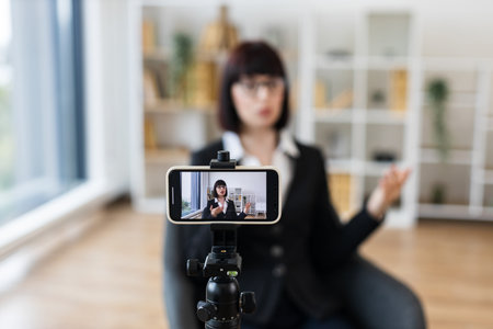 Caucasian woman recording video blog indoors with smartphone on tripod, sharing business insights and advice. She has dark hair, glasses, wearing formal clothing.の写真素材