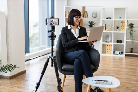 Young Caucasian woman in formal attire recording professional blog content on camera in spacious office environment, smiling while using laptop, showcasing modern working setting involving technologyの写真素材