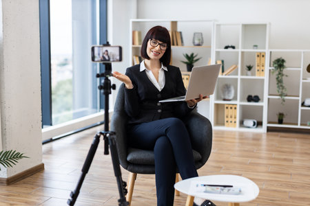 Young Caucasian woman in formal attire recording professional blog content on camera in spacious office environment, smiling while using laptop, showcasing modern working setting involving technologyの写真素材