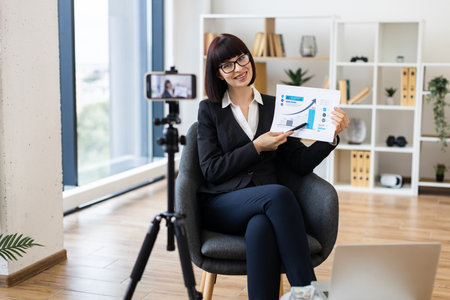 Caucasian woman presenting business analytics on camera through blogging using report and statistics chart.の写真素材