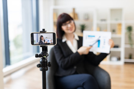 Caucasian woman in business suit recording video about revenue growth using graphs, modern office setup with natural light, professional presentation, concept communication and education.の写真素材