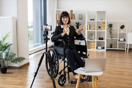 Caucasian adult woman seated in wheelchair recording online business content using smartphone and laptop. Modern office setting with shelves, indoor plants, and natural light.の写真素材