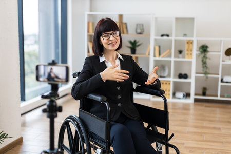 Caucasian woman wearing business attire sits in wheelchair recording video blog in office using smartphone. Professional setting conveys ideas of inclusivity accessibility business presentationの写真素材