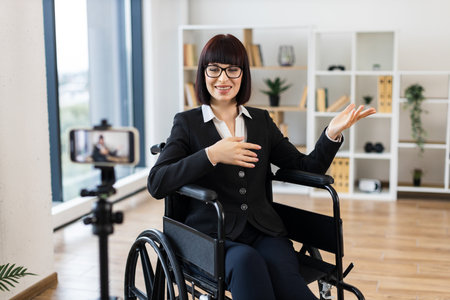 Caucasian woman wearing business attire sits in wheelchair recording video blog in office using smartphone. Professional setting conveys ideas of inclusivity accessibility business presentationの写真素材