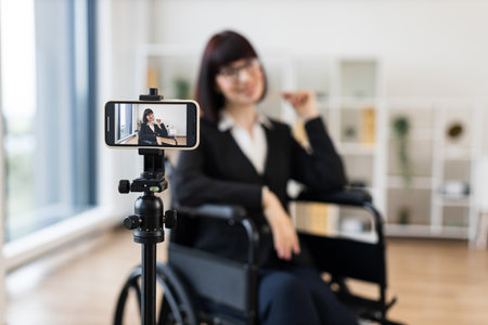Caucasian woman wearing formal clothes sits in wheelchair recording business blog onto camera at office. Motivational and entrepreneurial vibes emphasizing inclusion, diversity, professional insights.の写真素材