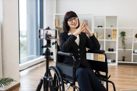 Caucasian woman wearing formal clothes sits in wheelchair recording business blog onto camera at office. Motivational and entrepreneurial vibes emphasizing inclusion, diversity, professional insights.の写真素材