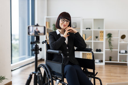 Caucasian woman wearing formal suit, sitting in wheelchair, recording business blog on smartphone camera in modern bright office. Represents entrepreneurship, determination, accessibility.の写真素材